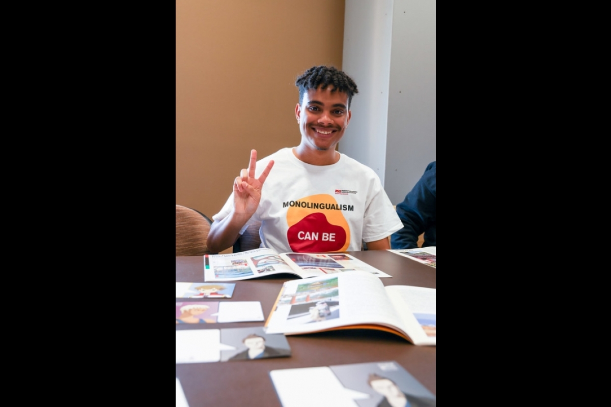 Person smiling giving a peace sign while tabling, wearing a 'monolingualism can be cured' shirt