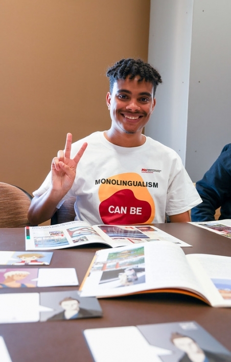 Person smiling giving a peace sign while tabling, wearing a 'monolingualism can be cured' shirt