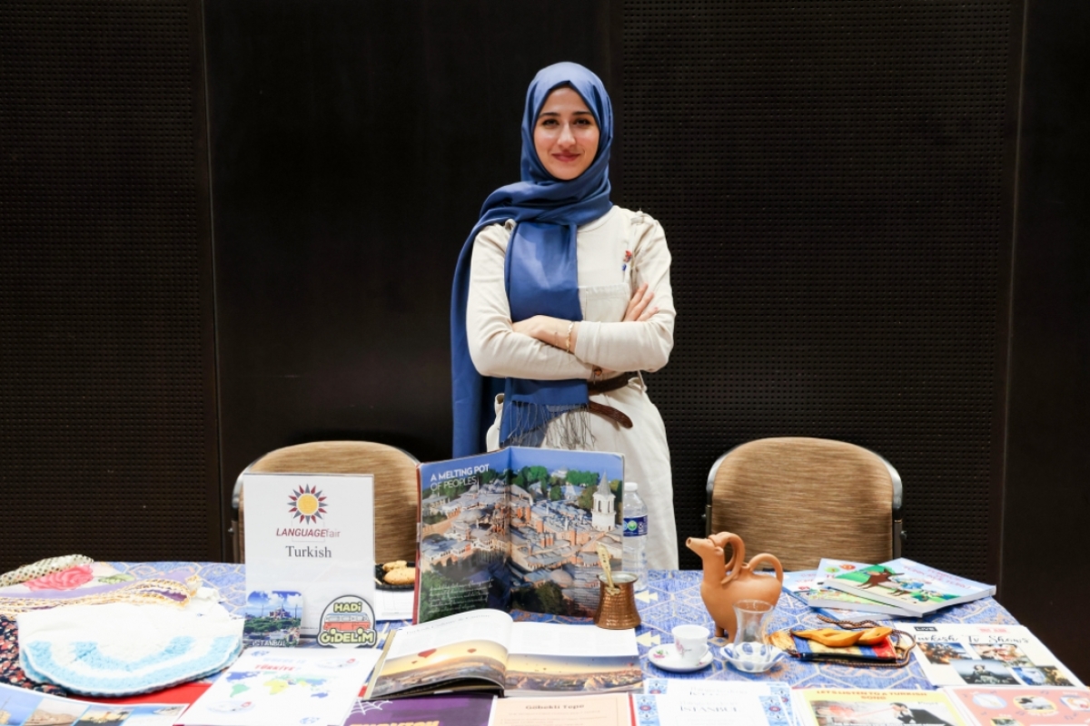 Smiling woman with arms crossed in front of the Turkish language table with various cultural items originating from Turkey resting on the table