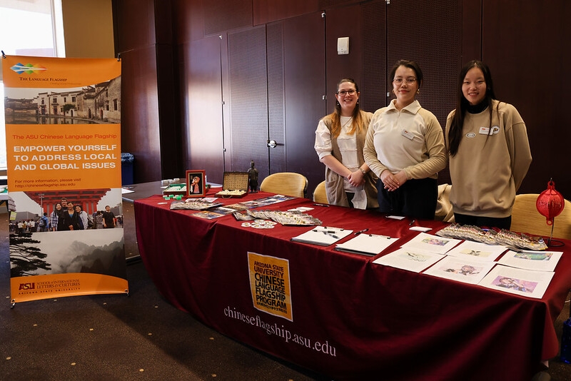 Three people standing behind a red table that the Chinese Language Flagship Program logo with various informative pamphlets on the table and a orange banner on the left of the table.