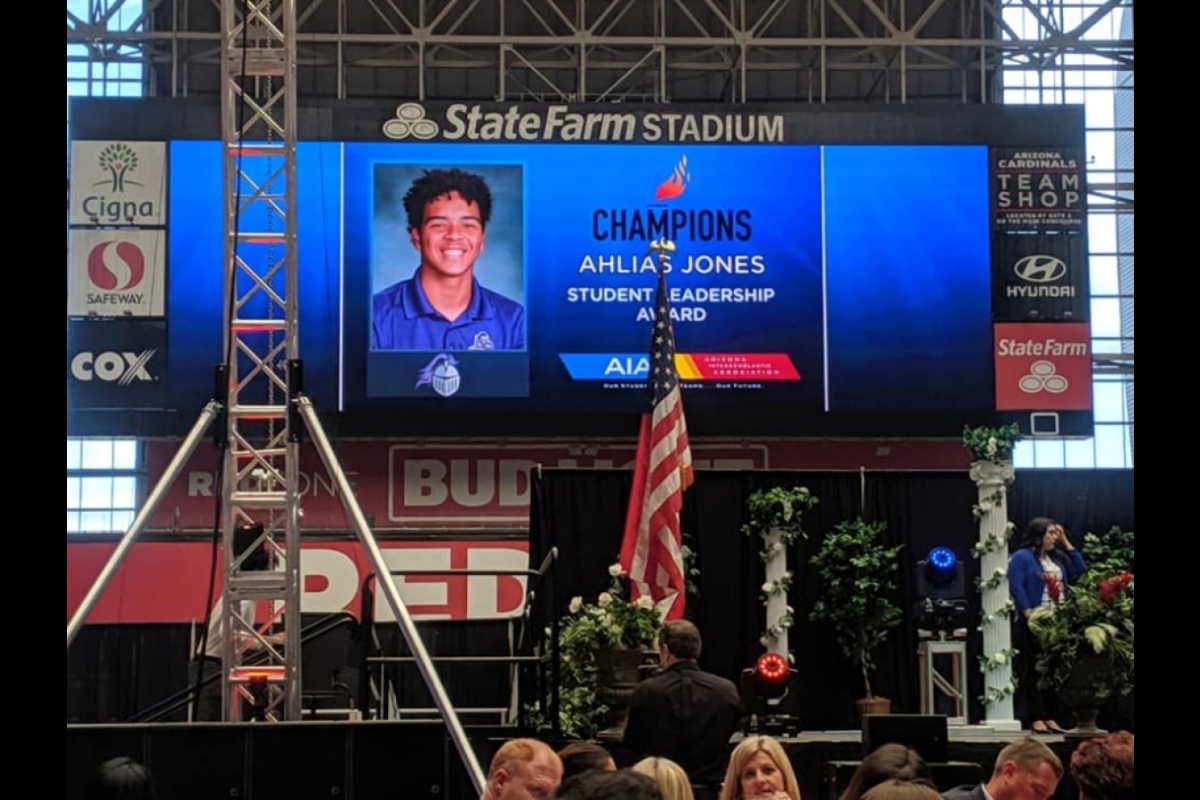 Large screen above a stage displaying a photo of a smiling young student identified as Ahlias Jones. Next to his photo are the words 