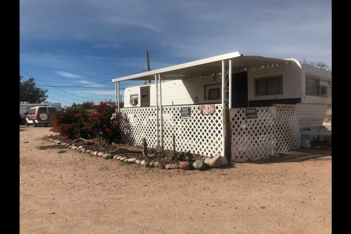 A white mobile home surrounded by a dirt lot and some bushes