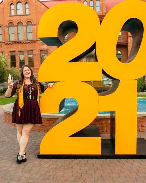 ASU College of Health Solutions graduate Emily Armstrong making the pitchfork sign with her hand as she poses next to a giant 2021 sign