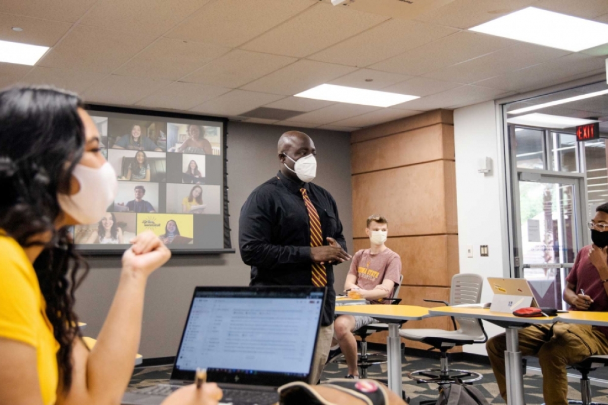 A professor in a mask speaks in front of a classroom of distanced, masked college students