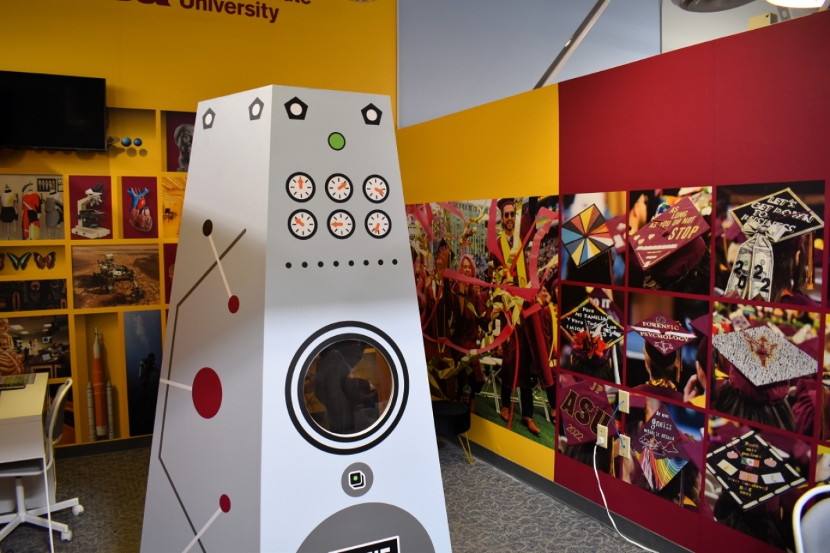 A mock machine with timers and levers stands in a space decorated with photos of graduating college students.