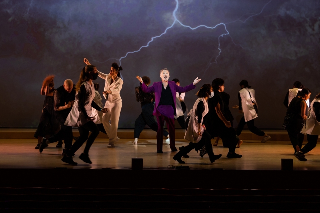 Man wearing a purple suit stands at the center of a stage while women wearing white and black walk around him and lightnight flashes in the background.