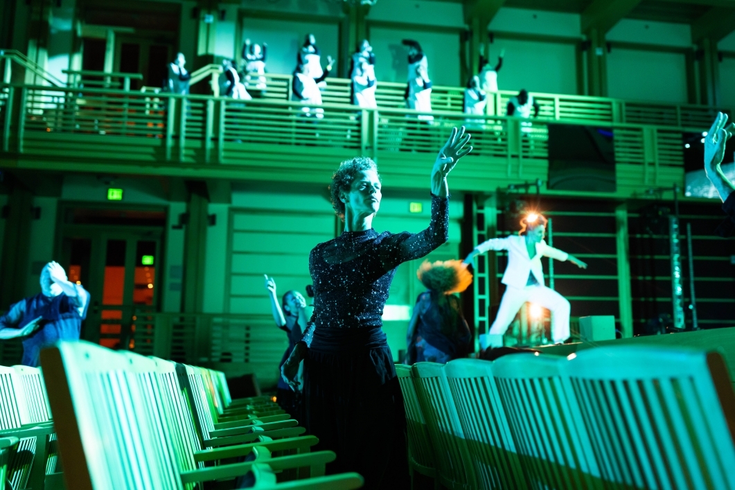 Women wearing black and white costumes move around an auditorium cast in a green light.