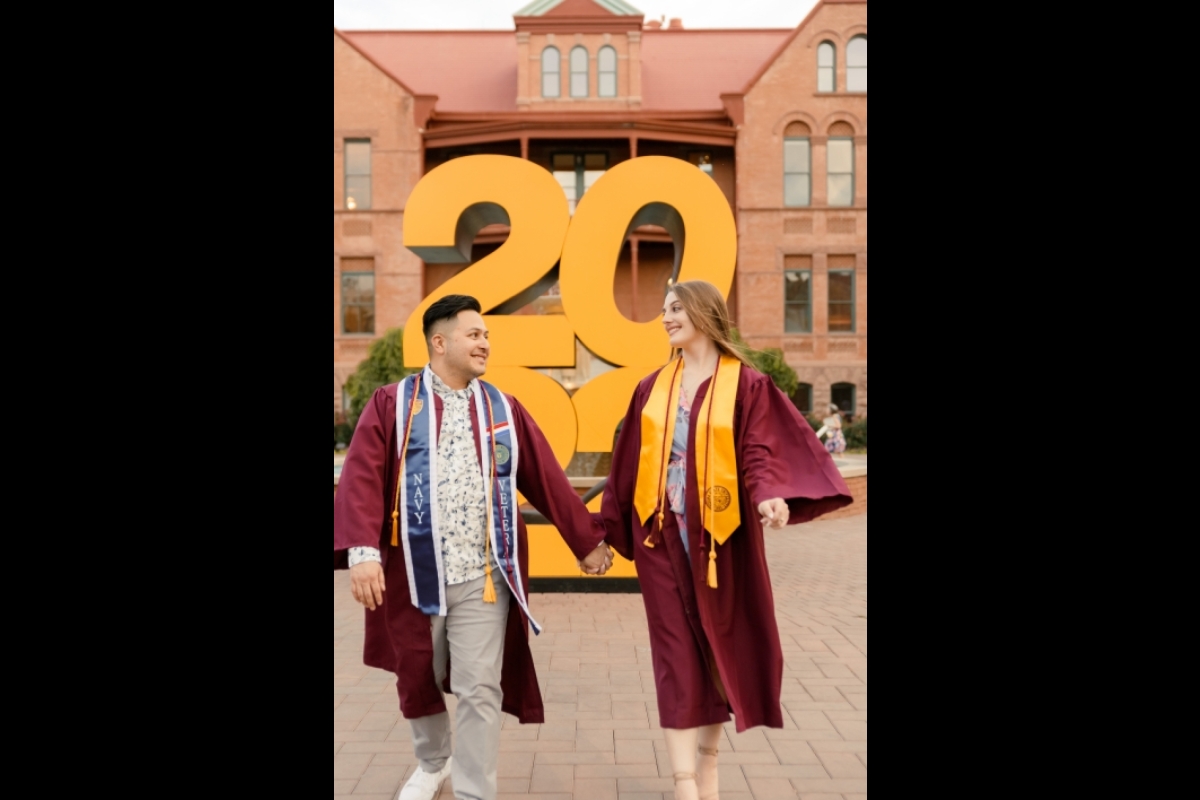 ASU graduates Alex Peraza and Chelsea Patti wearing graduation regalia and holding hands in front of a large 2022 sign.