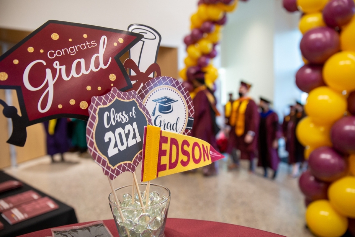 Signs saying "Congrats grad," "Class of 2021" and "Edson" are displayed in the foreground. In background is a maroon and gold balloon arch with graduates walking through it.