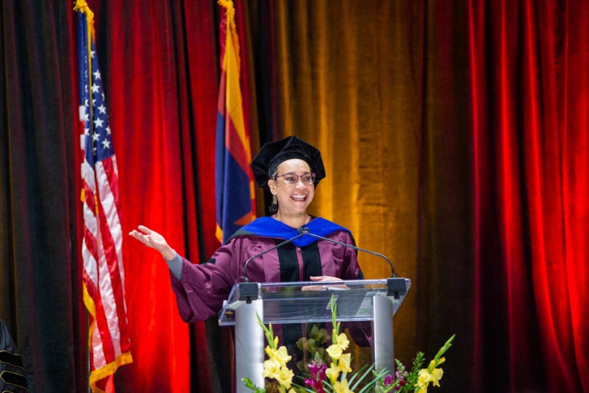 Kenja Hassan speaks at the podium wearing a maroon gown with a blue hood and black cap.