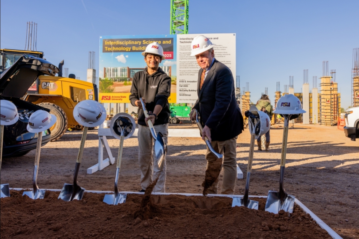 Two men wearing hard hats use ceremonial shovels to move dirt at a groundbreaking ceremony.