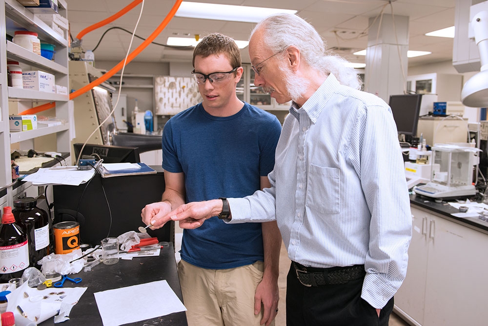 An older man with white hair in a ponytail speaks to a younger man in a T-shirt in a lab setting.