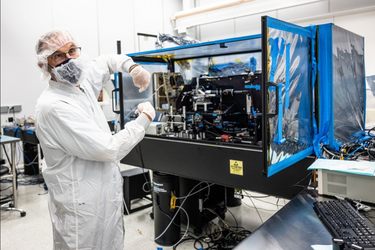 A man in a lab coat and hair nets points to a space camera in a clean room lab