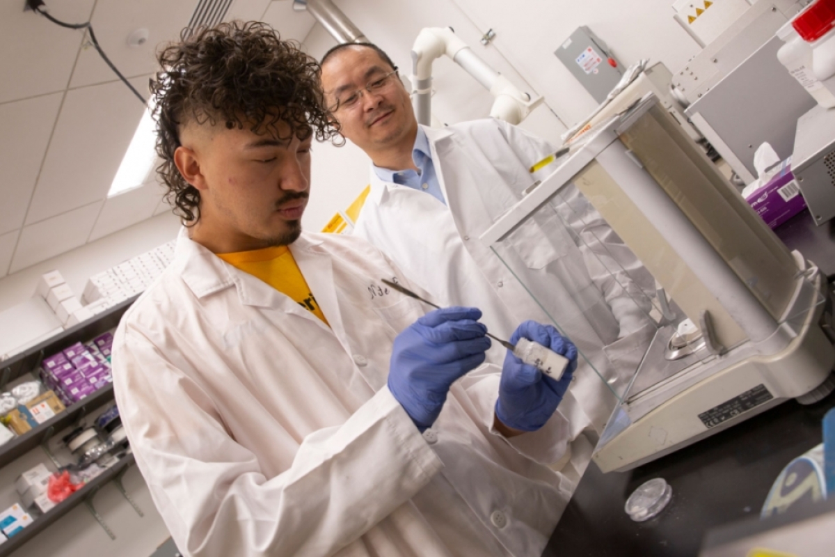 ASU student Nathan Fonseca wearing a white lab coat and gloves as he works with tools in a lab and ASU Assistant Professor Kenan Song looks on.