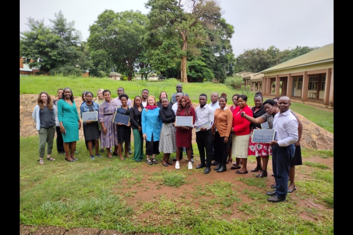 All the participants from the workshop in Lilongwe pose together for a group photo. They are outside in a grassy area with buildings behind them to one side and trees behind them to the other side.
