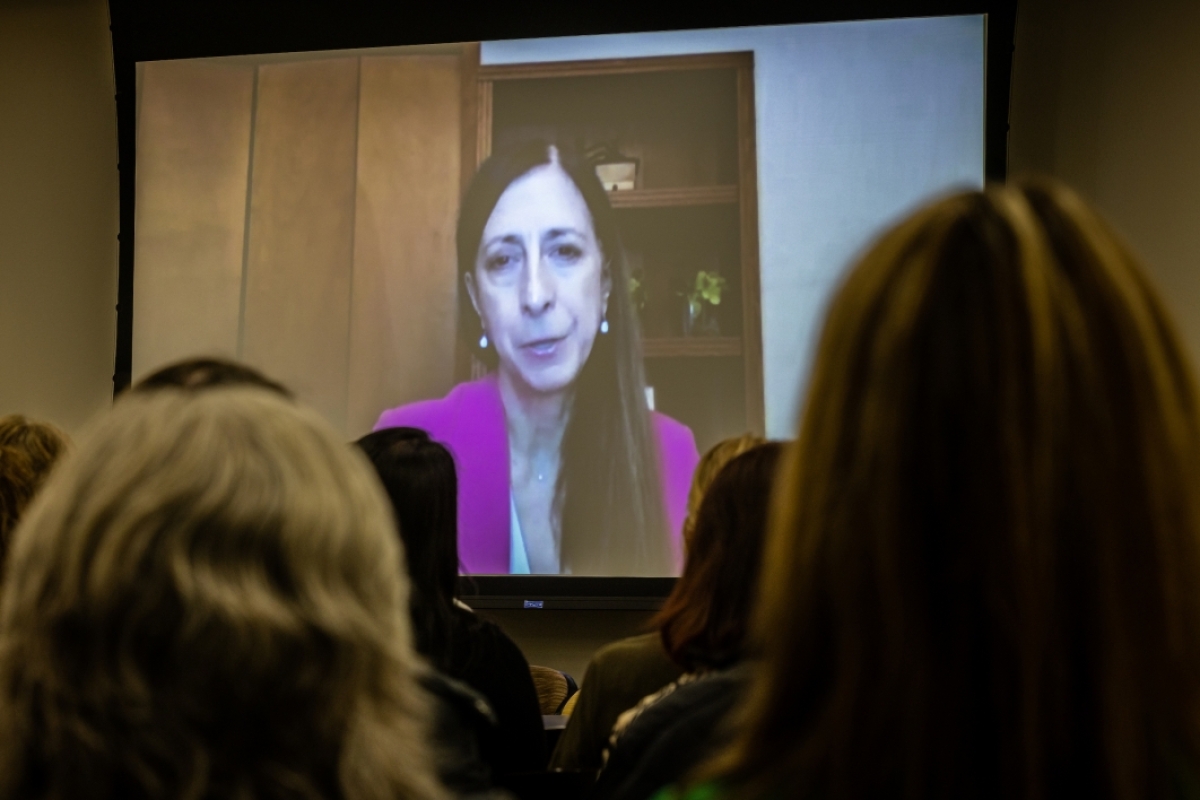 Woman's face projected on a screen as she speaks to a crowd via Zoom.