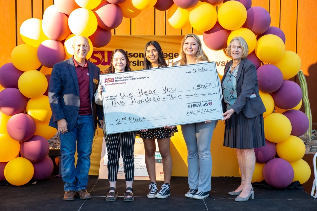 Three students hold a large check with on stage with a balloon backdrop behind them