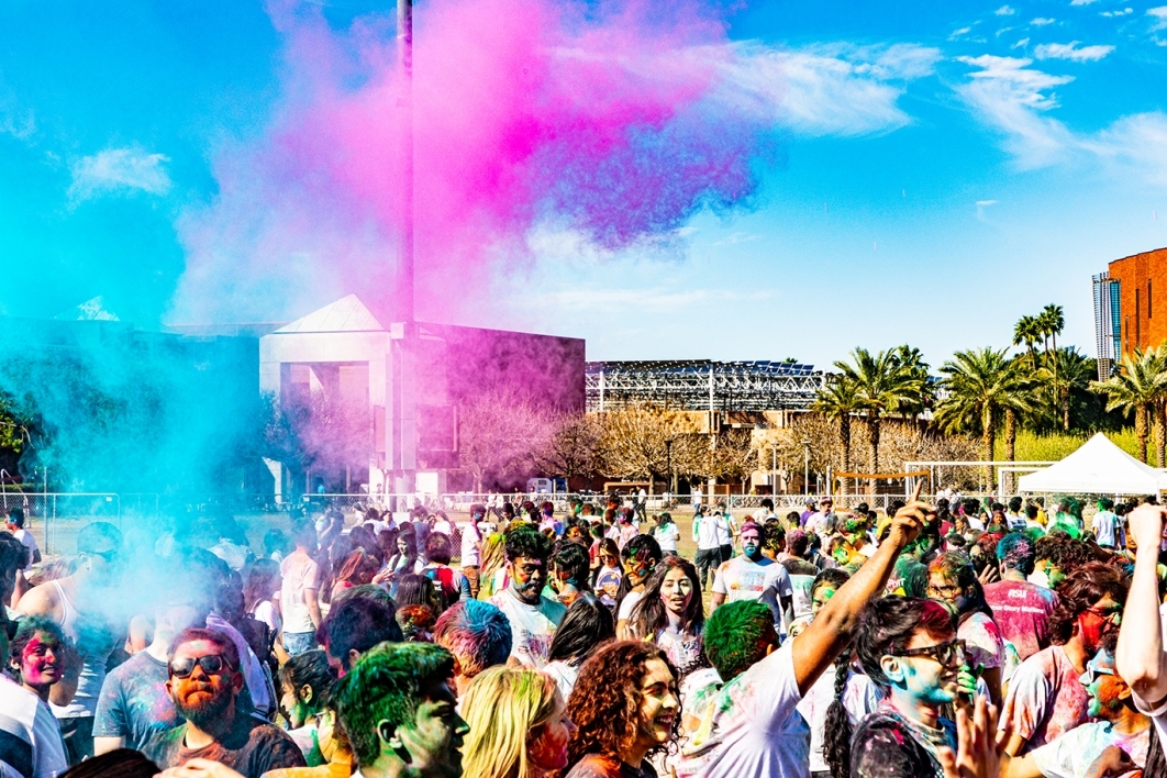 Group of Holi attendees under colorful puffs of powder in the air.