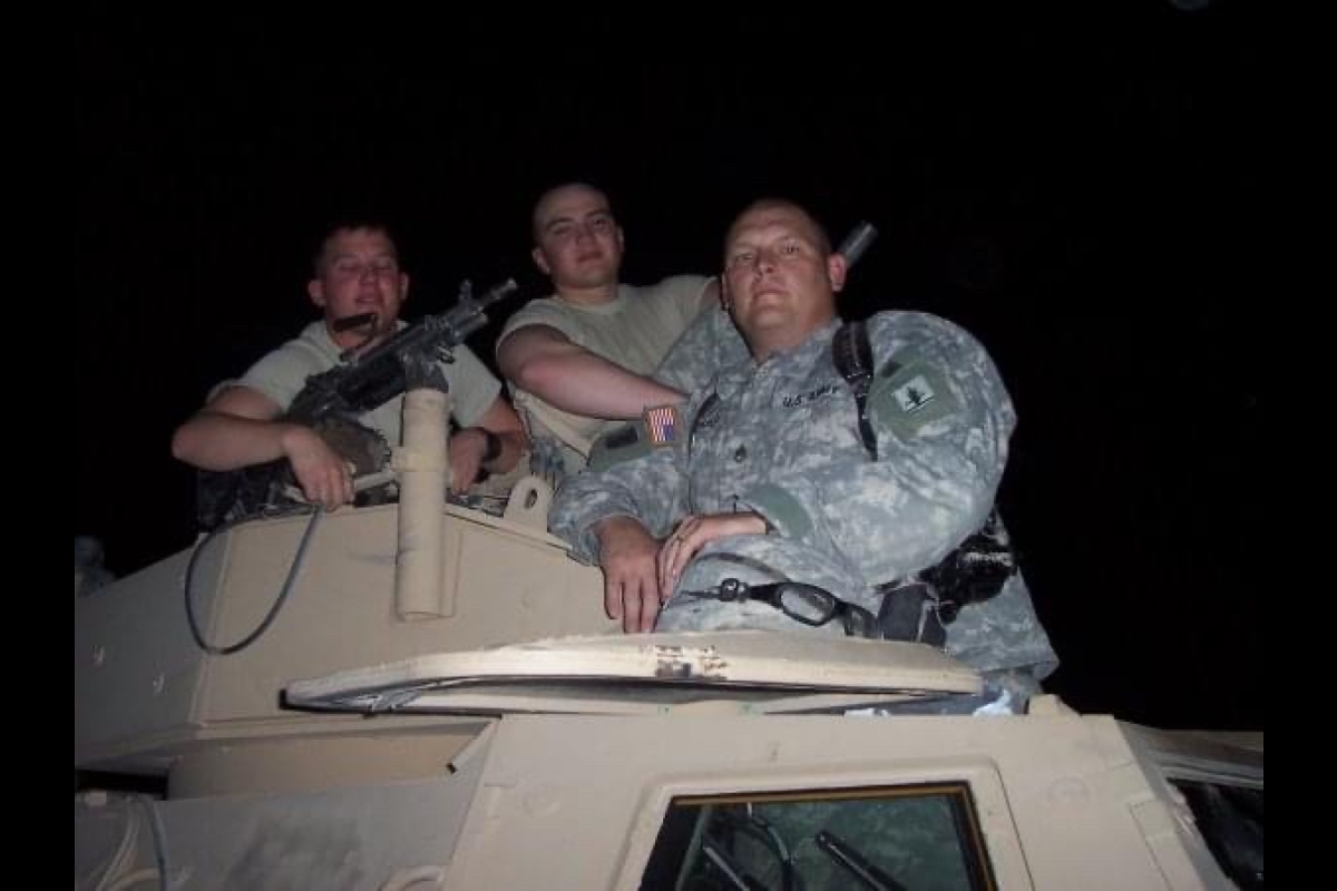 Men wearing military fatigues pose with large guns on the top of a military vehicle.