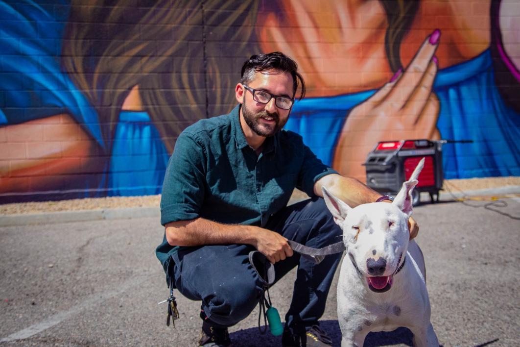 The artist, wearing glasses and a green button-up shirt, crouches on the pavement in front of the mural with his pit bull