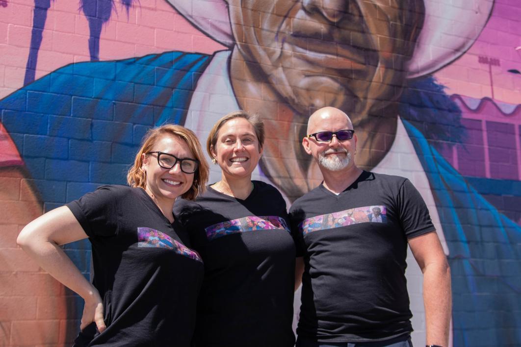 Two females and one male stand in front of the Maryvale mural wearing T-shirts also depicting the mural
