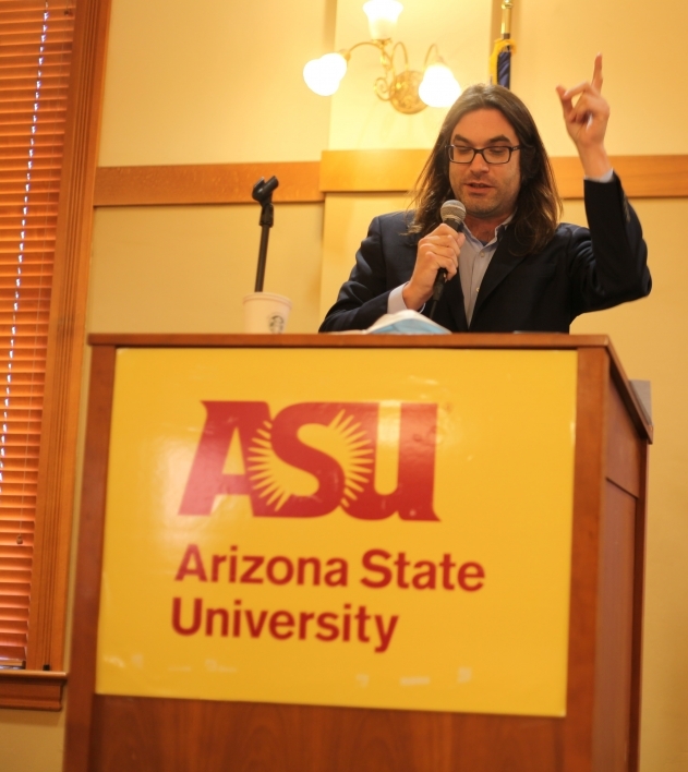 William Hedberg presents his research. He is wearing a dark suit coat over a lighter collared button-down shirt. His left hand is raised in the air as he talks with his index finger pointing upward. He stands at a wooden lectern with the ASU logo on it.
