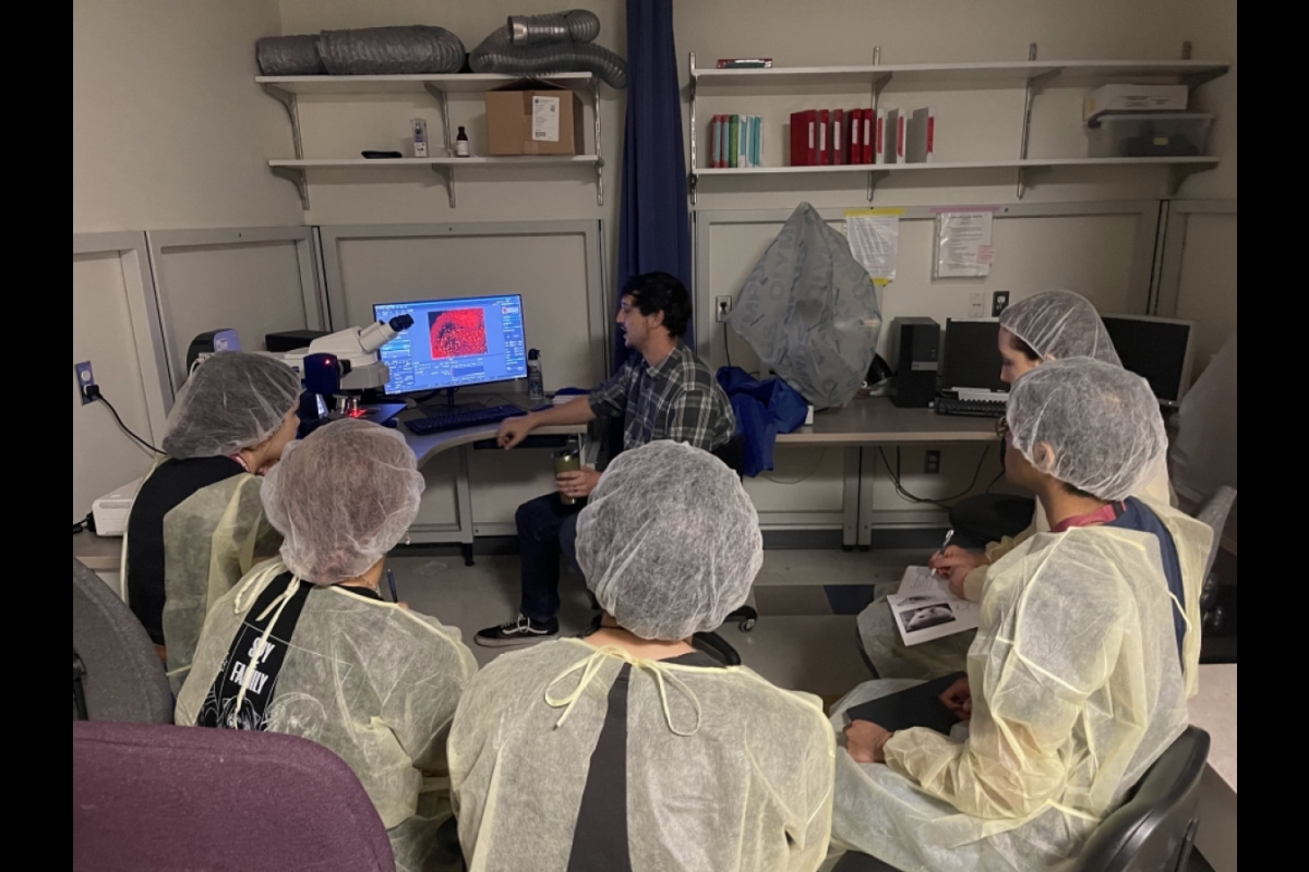 Graduate student seated at a desk with a computer and microscope talks to a group of high school students wearing surgical coverings.