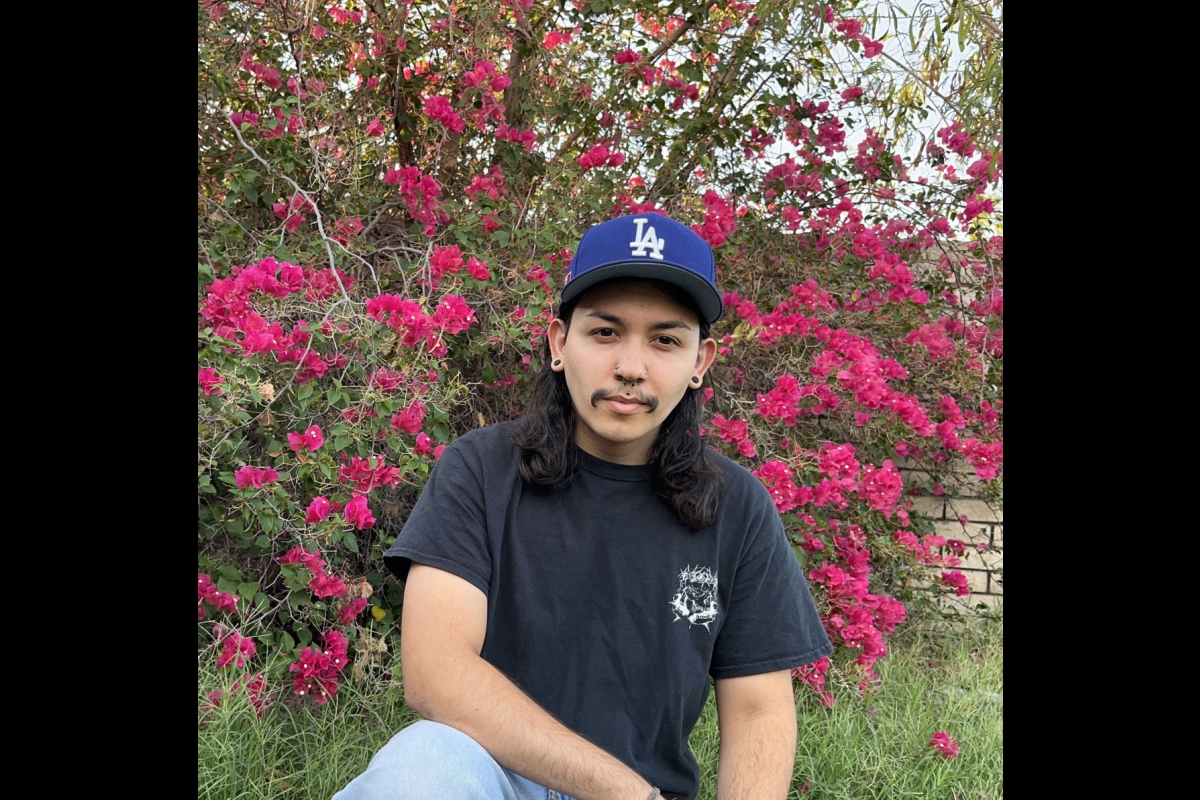 Man with shoulder-length black hair wearing a black T-shirt and jeans and an L.A. Dodgers cap sitting in front of a bougainvillea bush.