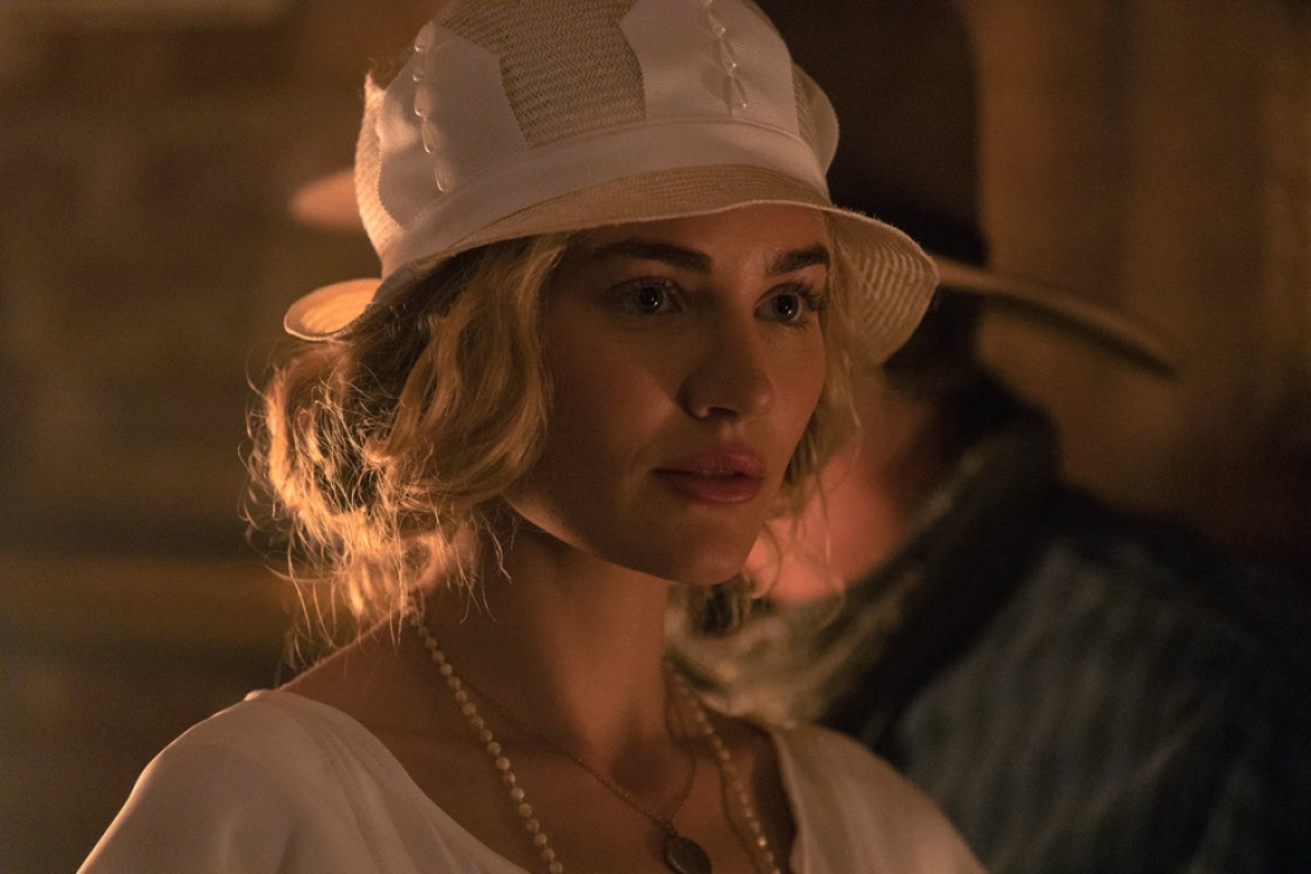 A blonde woman in a 1920s hat looks serious in a dimly lit interior.