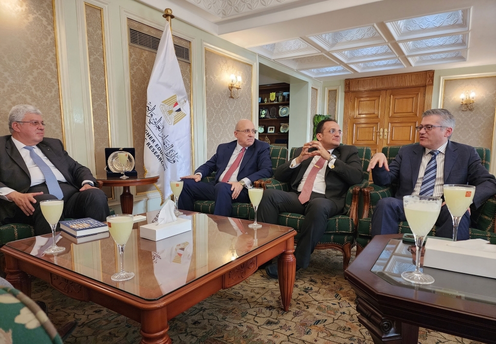 Four men in suits sit around a large coffee table with various flags behing them