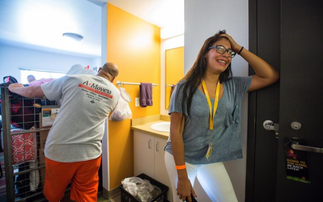 A student holds open her dorm door as movers take her stuff inside.