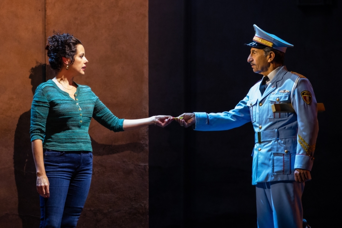 A woman wearing street clothes accepts a peice of paper from a man wearing a uniform in a scene from the play 