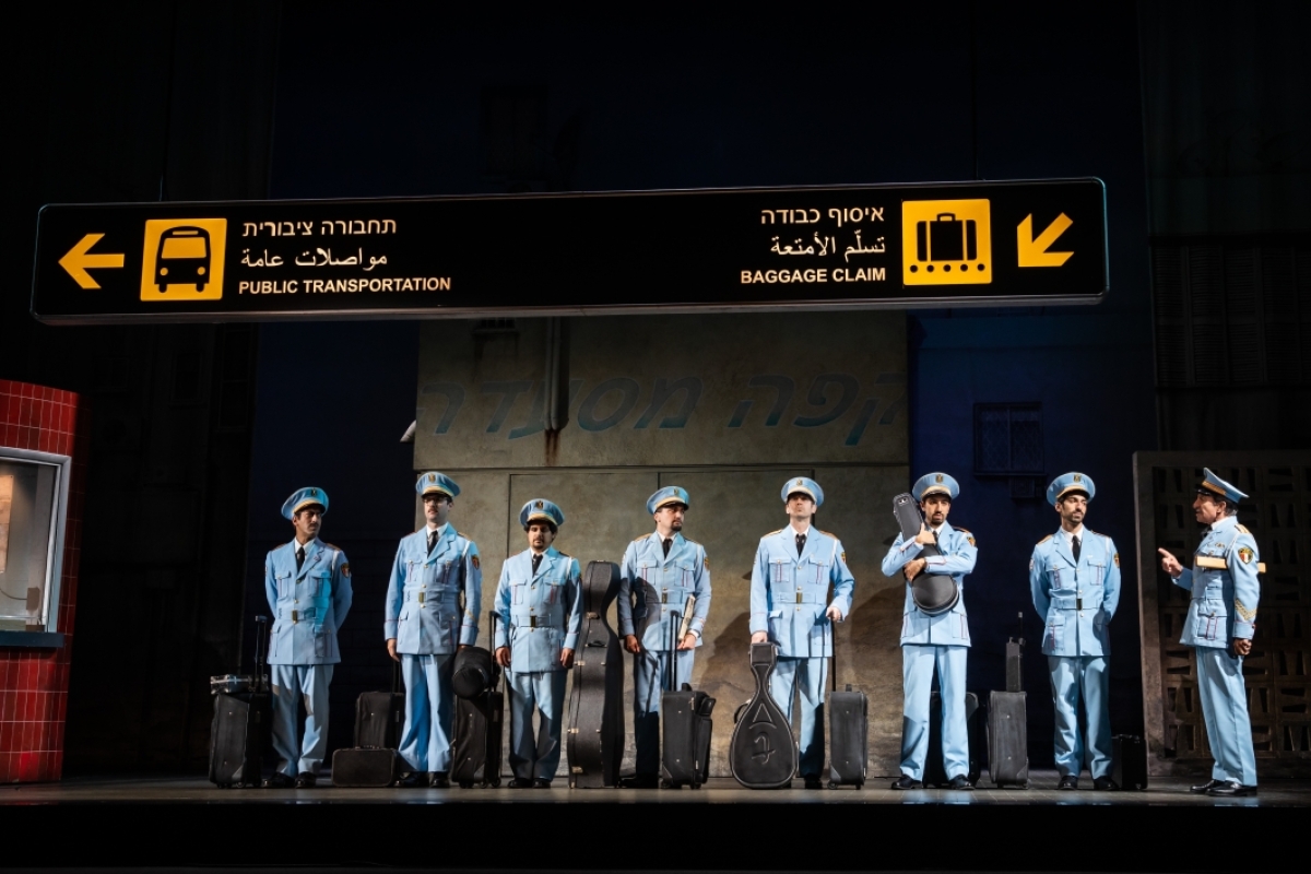 Men wearing uniforms and holding instrument cases stand in a row on stage in a scene from "The Band's Visit."
