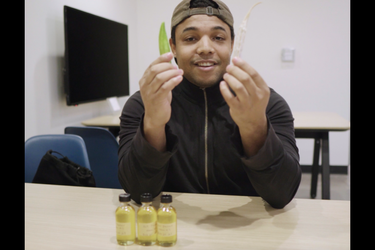 Jordan Collins holds holding an immature and mature okra pod with three small bottles of okra seed oil on table in foreground