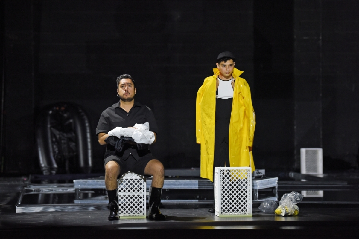 One seated man and one standing man, wearing a yellow raincoat, onstage.