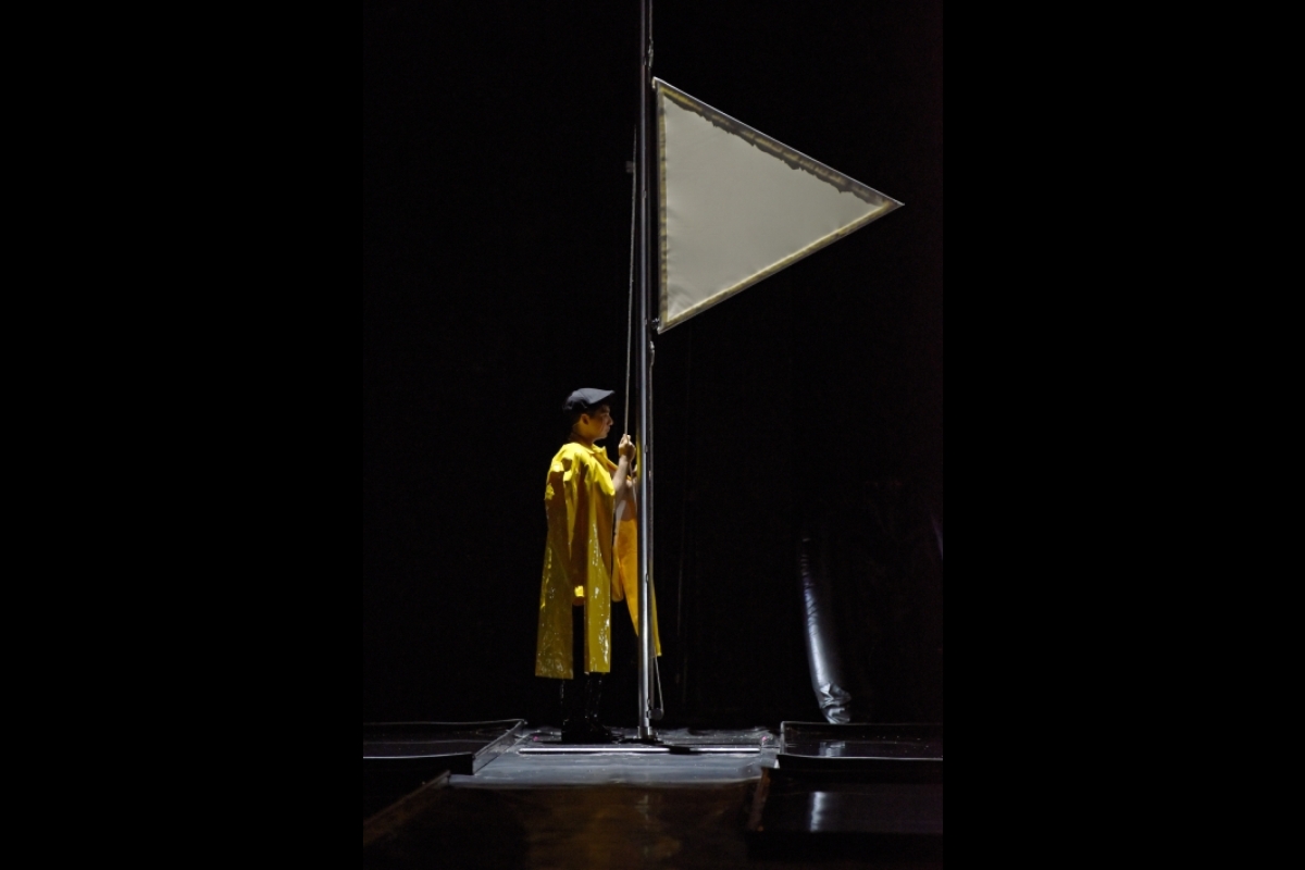 Man wearing a yellow raincoat standing in a boat with a sail onstage.