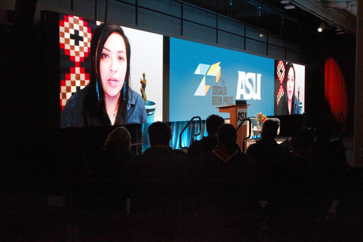 A woman on a large screen is presented to an audience.