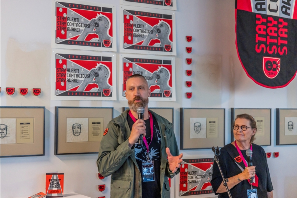 Man speaking into a microphone next to a woman holding a microphone in front of a wall displaying works of art.