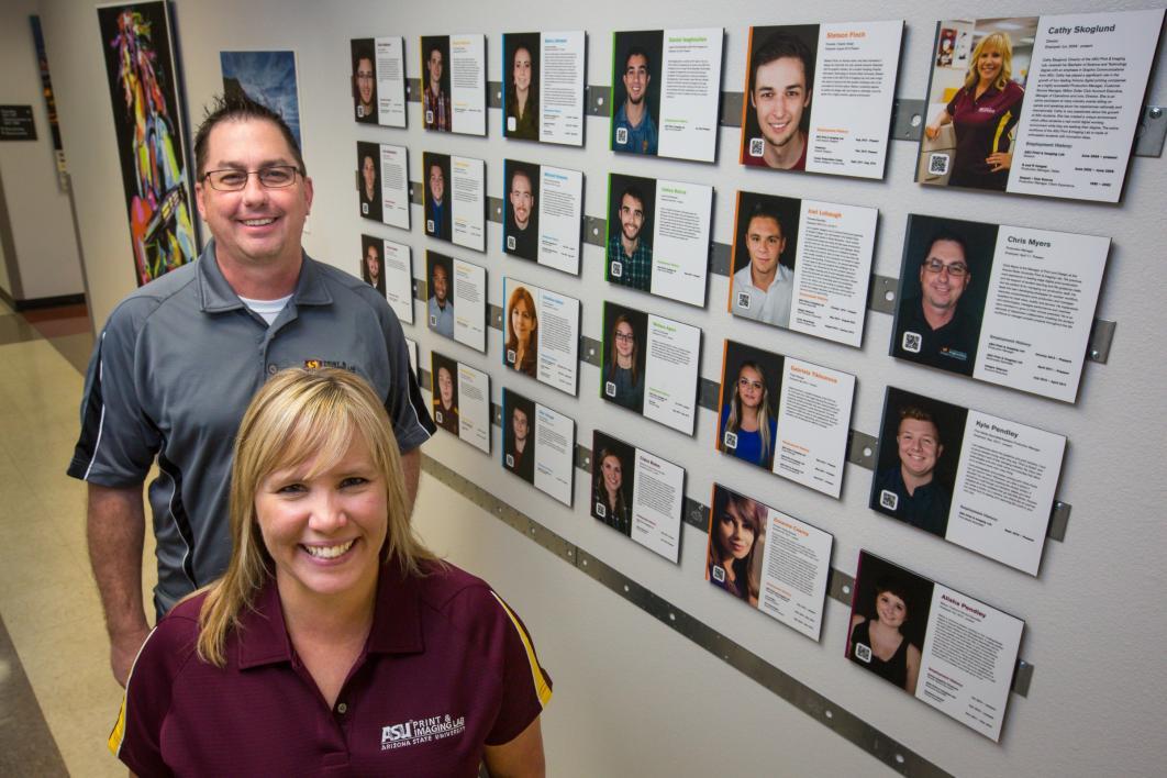 two ASU staff members posing next to wall of photos