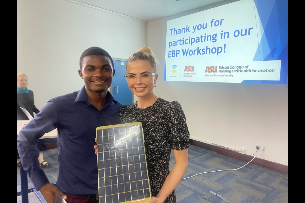 Rachel Thompson smiles at the camera with a workshop participant while holding a SolarSPELL device. There is a powerpoint slide in the background that says, "Thank you for participating in our EBP Workshop!"