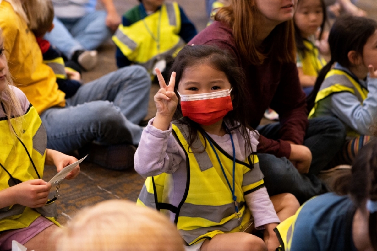 Child sitting in a group of other children and Mirabella at ASU residents, making a peace sign with her hand.