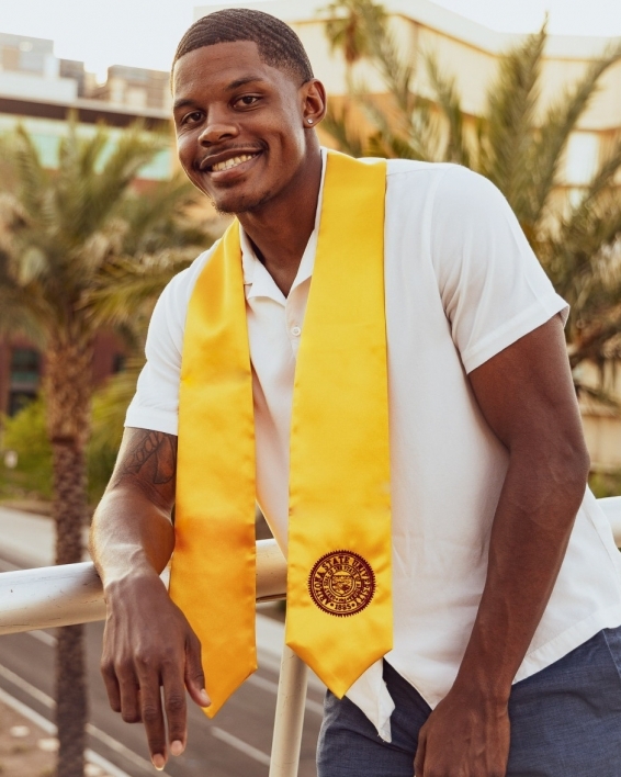 ASU's College of Health Solutions graduate James Rouser smiles while wearing his graduation sash in front of some palm trees