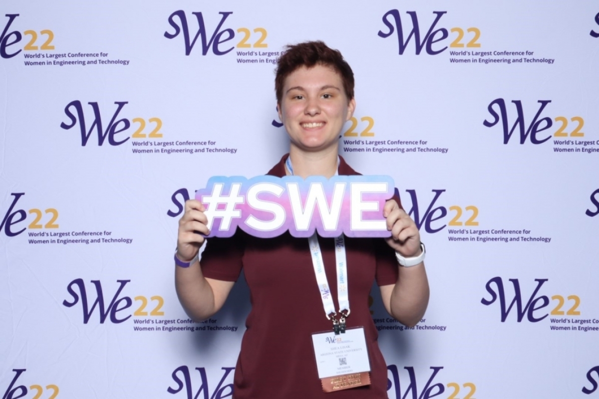 Woman standing in front of a wall with the logo "SWe22," holding a sign that reads "WE22."