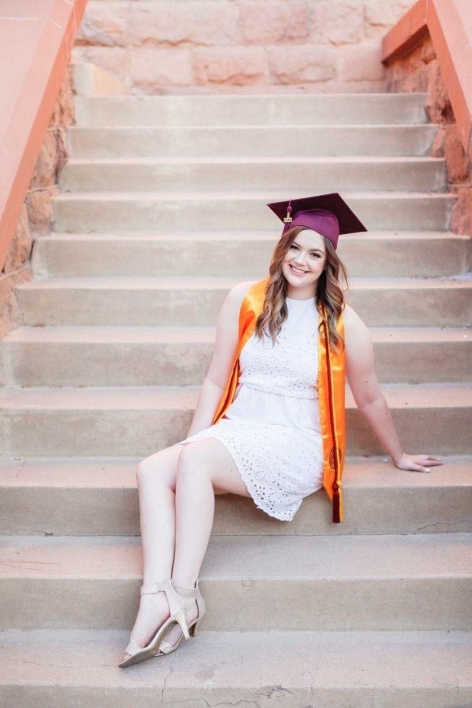 ASU's College of Health Solutions graduate Paige Snyder smiles in a white dress, wearing her graduation cap and sash while sitting on a staircase