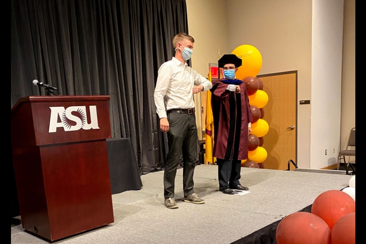 A student in PhD commencement robes bumps elbows with his adviser on stage