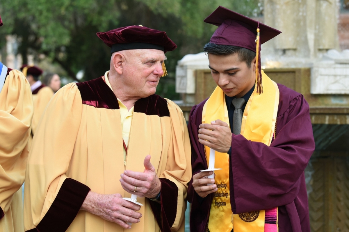 An older man leans toward a new graduate, both in graduation regalia, as they light candles