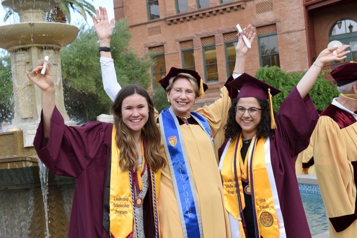 Two new grads in maroon regalia throw their arms up to celebrate in a photo with a 1972 grad in golden robes