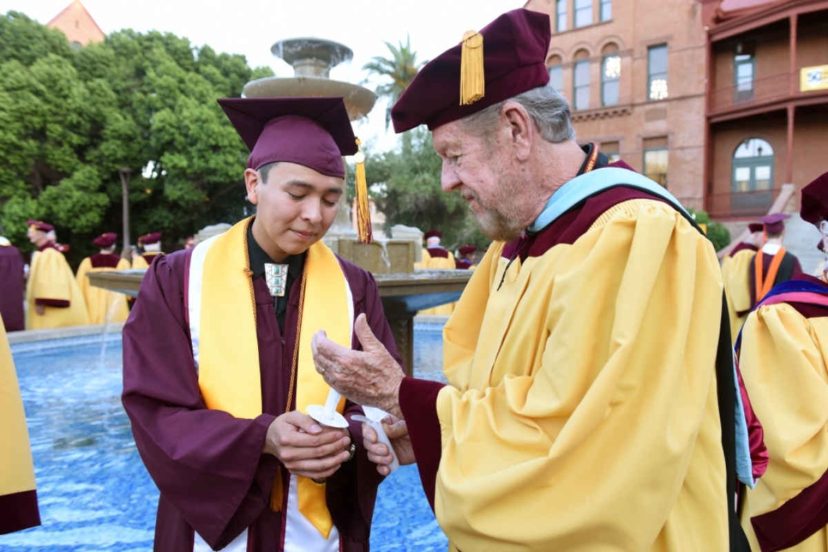 Two Sun Devils passing the flame from candle to candle wearing graduation gowns and caps.