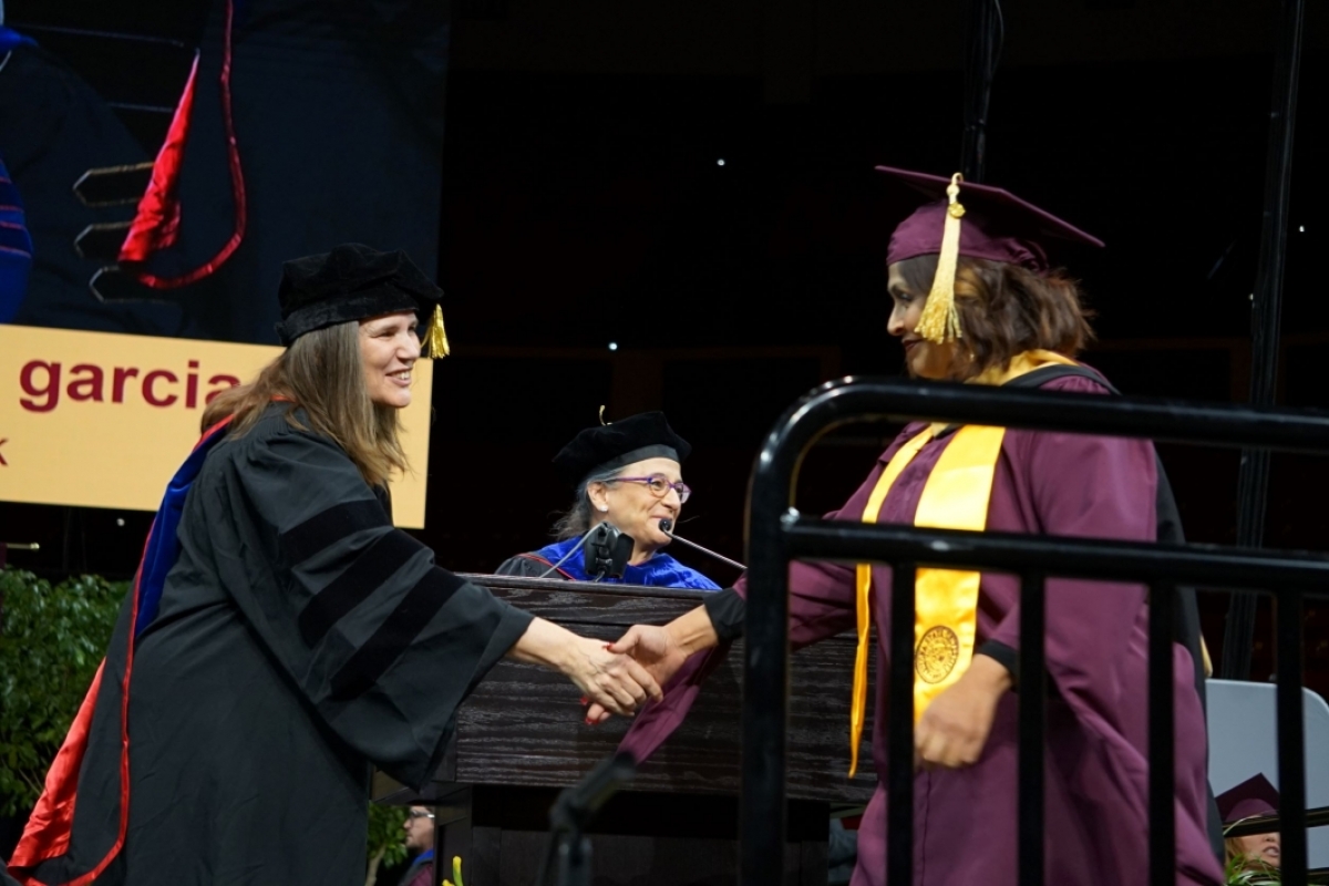 Two women wearing graduation regalia and shaking hands on a stage.