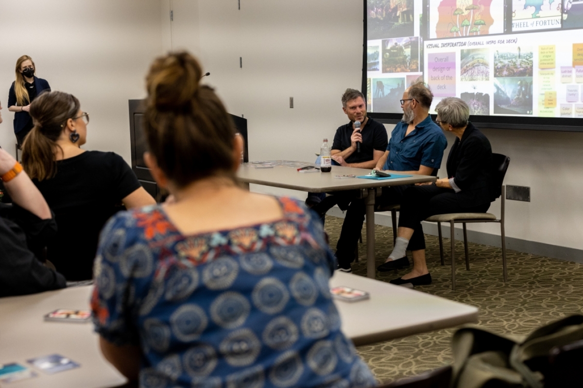 Photograph of three panelists (Neil Smith, Dr. Gaymon Bennett, and Dr. Elizabeth Langland) speaking at an event, with several audience members looking on in the foreground. 